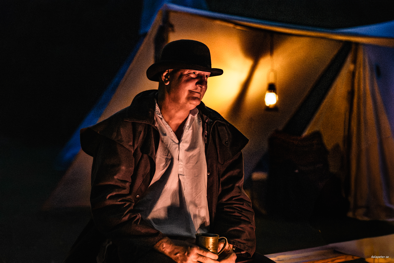 Stämningsfullt och filmiskt porträtt av en man i plommonstop vid en lägereld utanför ett tält under den blå timmen. Fotograf: Peter Gunnebro
